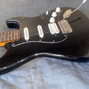 Černá elektrická kytara typu Stratocaster s bílým pickguardem a třemi bílými snímači. Nástroj má tři bílé oválné ovládací knofíky a jeden bílý přepín…