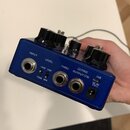 Modrý kytarový efekt s černými oválnými knoflíky. Na zadní straně jsou konektory a nápisy INPUT, LEVEL, THRU, (TRS) OUTPUT (TS), USB a DC 9V. U přepí…