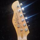 Fotografie zachycuje hlavu elektrické kytary z přírodního světlého dřeva s šesti chromovanými ladičkami. Na povrchu je černý nápis „Customman GUITARS…