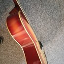 Detailní pohled na tělo polodupaté elektrické kytary s červeným sunburst povrchem a krémovým lemováním. Na těle je vidět jeden f-otvor a část hmatník…