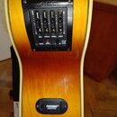 Detailní pohled na ovládací panel akustické kytary s žlutým přechodem do oranžové barvy. Černý panel nese nápis AEQ a obsahuje čtyři posuvníky s popi…