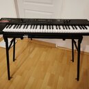 Černý digitální klavír umístěný na černém X-stojanu. Na levé straně korpusu je bílý nápis Roland a pod ním modelové označení Juno-D, doplněný červený…