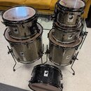 Sada bubnů v šedé metalické barvě s černými obručemi a kováním. Na korusech jsou viditelná loga „Helen Drums“. Soubor obsahuje pět bubnů různých veli…