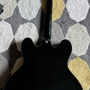 Zadní strana polodubové elektrické kytary v černé barvě s lesklým povrchem. Na hlavě nástroje je vidět nápis Epiphone a šest zlatých ladiček. Kytara…