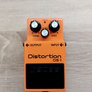 Oranžový efekt pedál s nápisy Distortion a DS-1. Na horní části jsou tři ovládací knoflíky s popisky TONE, LEVEL a DIST a červená kontrolka s nápisem…