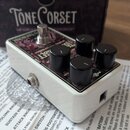 Bílý efekt pedál s černými ovládacími knoflíky a stříbrným spínacím tlačítkem. Na horní ploše je fialový grafický panel s nápisy Tone Corset a Compre…