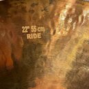 Detailní záběr na povrch mosazného činele. Na povrchu je vyražen nápis 22" 55 cm RIDE. Povrch vykazuje výrazné oxidace, tmavé skvrny a známky opotřeb…
