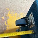 Detailní záběr na kovové kolečko s modrou gumovou pláští, které je přišroubované k tmavě modrému plstnatému povrchu. Vedle kolečka je přiložen žlutý…