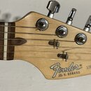 Detail hlavy světlé dřevěné elektrické kytary s chromovanými ladičkami. Na hlavě je černé logo Fender a nápisy STRATOCASTER, MADE IN U.S.A. oraz SERI…