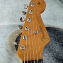 Fotografie zachycuje hlavu elektrické kytary z přírodního dřeva. Na povrchu je vidět logo Fender a nápis STRATOCASTER, pod kterým je umístěn menší, n…
