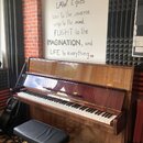 Hnědé dřevěné digitální piano s lesklým povrchem a černou klaviaturou. Na přední liště pod víkem je zlatý nápis PELTROE. K nástroji patří černá polst…
