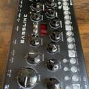 Černý syntezátor s nápisem BASSLINE a označením DB-01 v pravém dolním rohu. Na horní hraně je logo ercosynths. Přístroj obsahuje několik černých otoč…