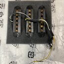 Tři černé elektrické snímače typu humbucker umístěné na šedé pěnové podložce. Snímače mají viditelné měděné cívky a jsou připojeny k bílým, žlutým a…