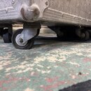 Detail spodní části stříbrného transportního kufru z hliníku s kovovými hranami a rohy. Kufr je osazen čtyřmi černými kolečky na kovových držácích. P…