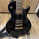 Černá elektrická kytara s gold hardwarem a bílým lemováním korpusu. Na horní části korpusu jsou nápisy "RHYTHM" a "TREBLE". Nástroj má dva humbuckery…