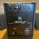 Zadní strana černého aktivního reproduktoru značky HK AUDIO, model LUCAS XT. V horní části jsou vstupy a výstupy s popisky MIC IN, LINE IN, AUX IN a…