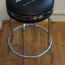 Černý kulatý taburet s chromovanou kovovou konstrukcí a kruhovou opěrkou pro nohy. Na horní části sedáku je bílý nápis „Fender Custom Shop“ a pod ním…