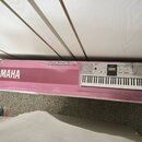 Fotografie zachycuje kartonovou krabici od klávesového nástroje v růžové a bílé barvě. Na krabici je nápis YAMAHA s logem značky a označení modelu PS…