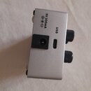 Stříbrný kovový box s dvěma černými knoflíky na pravé straně. Na horní části je nápis 9V 300mA doprovázený symbolem polarity a vedle něj nápis USB u…