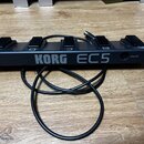 Černý pětitlačítkový pedál s nápisem KORG EC5. Tlačítka jsou od levého okraje k pravému označena písmeny E, D, C, B a A. V pravé části je vstup s náp…