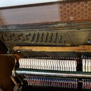 Detailní pohled do otevřeného vnitřku staršího klaviru. Je vidět kovový rám s ladicími kolíky, řada bílých klavišů a mechanika s červenými plstěmi. P…