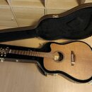 Akustická kytara s cutawayem v přirozené dřevěné barvě, umístěná v černém tvrdém kufru. Na hlavě hmatníku je viditelné logo „Takamine“. Povrch nástro…