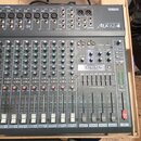 Šedý mixovací pult umístěný v dřevěném přepravním pouzdře. Na spodní části je vidět nápis YAMAHA a modelové označení Mx12/4. Zařízení obsahuje řadu p…