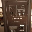 Zadní strana studiového monitoru v tmavě hnědé barvě. Na zadním panelu je bílý nápis FOCAL, ALPHA 65 EVO a PROFESSIONAL MONITORING SYSTEM. Jsou zde v…