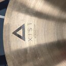 Detailní záběr na kovový činel zlatavé barvy s jemným broušením povrchu. Na povrchu je černý trojúhelníkový symbol a nápisy XIST a 18" Brilliant Cras…