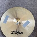 Kruhový činel z mosazi s viditelnými stopami po používání a dvěma šedými lepicími páskami na povrchu. Ve spodní části je černé logo Zildjian. U střed…