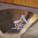 Detail zlatavého kovového činele s nápisy. Na povrchu je bílé logo ve tvaru stylizovaného písmene A a text CUSTOM 20th Anniversary Ride MEDIUM THIN 2…
