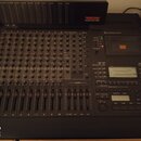 Černý mixážní pult s řadou potenciometrů a posuvníků. V pravé horní části je displej a ovládací sekce s nápisy jako SHUTTLE, MONITOR MODE a RECORD. V…
