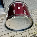 Basový buben v tmavě červené barvě s kovovým kováním. Povrch nástroje vykazuje viditelné opotřebení, škrábance a několik světlých skvrn. Na přední st…