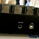 Detail zadní strany černého elektronického hudebního zařízení. Jsou vidět čtyři černé oválné ovládací knoflíky a dva konektory s nápisy SUSTAIN a USB…