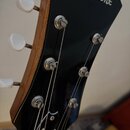 Detail hlavy černé elektrické kytary s bílým nápisem Epiphone. Na černé ploše jsou vidět kovové ladičky a stříbrné struny. Na hraně hlavy je vidět dř…