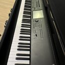 Černý digitální klavír v černém transportním kufru. Na pravé straně je bílý nápis KRONOS a pod ním menší text MUSIC WORKSTATION. Nástroj má displej,…