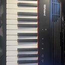Černý digitální klavír s bílými a černými klávesami. Na horním panelu je bílé logo Roland a napravo od něj kulaté tlačítko s písmenem G. Pod logem se…
