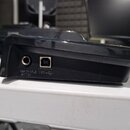 Detail zadní strany černého plastového zařízení. Jsou vidět dva konektory s nápisy ASSIGNABLE SWITCH a USB s ikonou USB. Povrch plastu je lesklý.