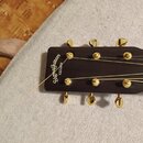 Detail hlavy akustické kytary v tmavě hnědé barvě se zlatými mechanikami. Na hlavě je zlatý nápis SigmaGuitars a pod ním menší text EST.1980. Povrch…