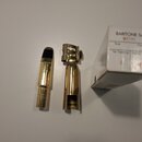 Kovový mouthpiece pro barytonsaxofon v zlaté barvě s černým ligaturem a bílou krabičkou. Na mouthpiece je vyryto logo "K 取り" a pod ním nečitelný text…
