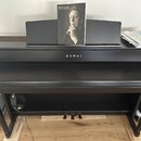 Černý digitální piano s uzavřenou klaviaturní víkem a integrovaným stojanem. Na přední straně je zlatý nápis KAWAI. Na notovém pultu je umístěn album…
