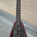 Elektrická kytara s V-tvarým korpusem v tmavě červené barvě s viditelnou strukturou dřeva. Nástroj má černý plech, dva humbuckery a tři ovládací knof…