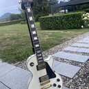 Bílá elektrická kytara s černým hmatníkem a černým pickguardem, umístěná na stojanu. Na hlavě hmatníku je zlaté logo Gibson. Nástroj má dva humbucker…