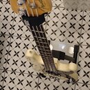 Krémově bílá elektrická basová kytara s jasným lakem a hnědým hmatníkem. Na hlavě nástroje je viditelné logo Fender. Kytara leží na digitální váze, k…
