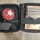 V otevřeném černém pouzdře je umístěna kalimba zC světlého dřeva s kovovými plátky. Na spodní části nástroje je vyrytý lineární obrázek a v horní čás…
