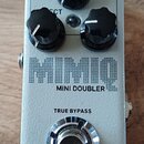 Stříbrný efekt pedál s třemi černými oválnými knoflíky. Na povrchu je nápis MIMIQ, MINI DOUBLER a TRUE BYPASS. Ve spodní části je umístěno logo a náp…