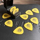 Žluté kytarové trsátka s nápisem DUNLOP U.S.A. 73 mm a doprovodným logem. Část trsátek leží volně na černém povrchu, dvě jsou umístěna v malém černém…