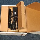 V otevřené kartonové krabici s nápisem Portico je umístěno černé elektronické zařízení a napájecí kabel. Na boku přístroje je svisle napsané logo nev…