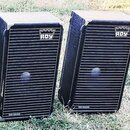 Dva černé reproduktorové boxy s hranatým tvarem a kovovými rohy. Na přední mřížce je v horní části bílé logo s nápisem ROY a v dolní části nápis TOP…