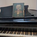 Černý pianino s klaviaturou. Na vnitřní straně víka nad klávesami je vyražen nápis KURZWEIHL. Na notovém pultu leží stará kniha s nápisy SANG u. KLAN…